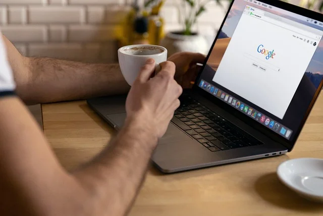 Professional sitting at desk with laptop showing Google search, holding coffee cup in modern workspace with brick wall background, representing strategic SEO research and link building expertise
